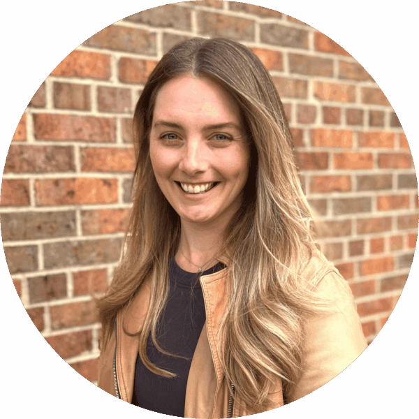 Smiling woman with long, light brown hair wearing a tan jacket and dark top, standing in front of a brick wall.