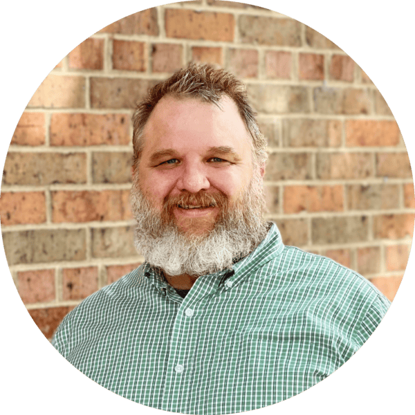 A man with a gray beard and short hair smiles at the camera, wearing a green plaid shirt. He stands in front of a brick wall. The photo is cropped in a circular frame.