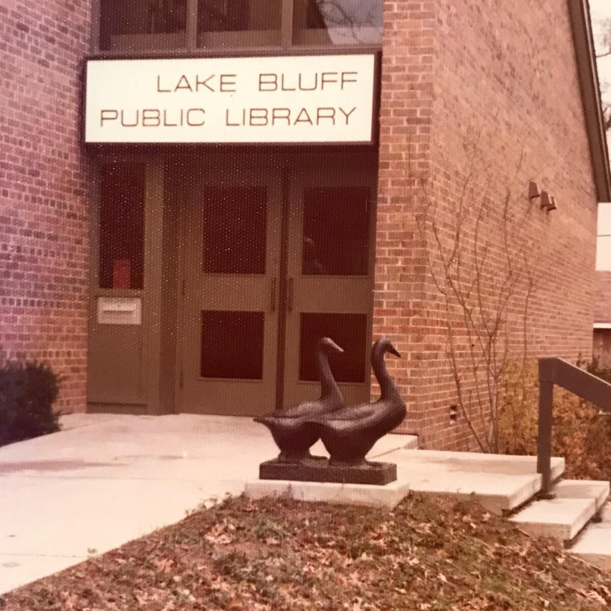 Entrance of Lake Bluff Public Library with a glass door, large sign above, and a sculpture of two geese on the sidewalk near brick walls and some bushes.