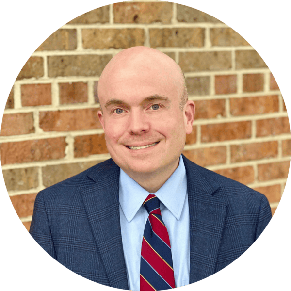 A man in a blue suit, light blue shirt, and striped tie smiles in front of a brick wall.