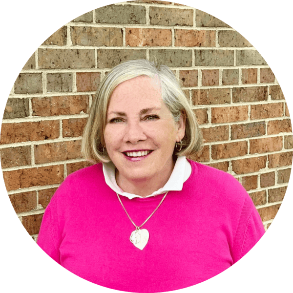 Smiling woman with gray hair in a pink sweater and white collared shirt, wearing a heart-shaped necklace, standing in front of a brick wall.