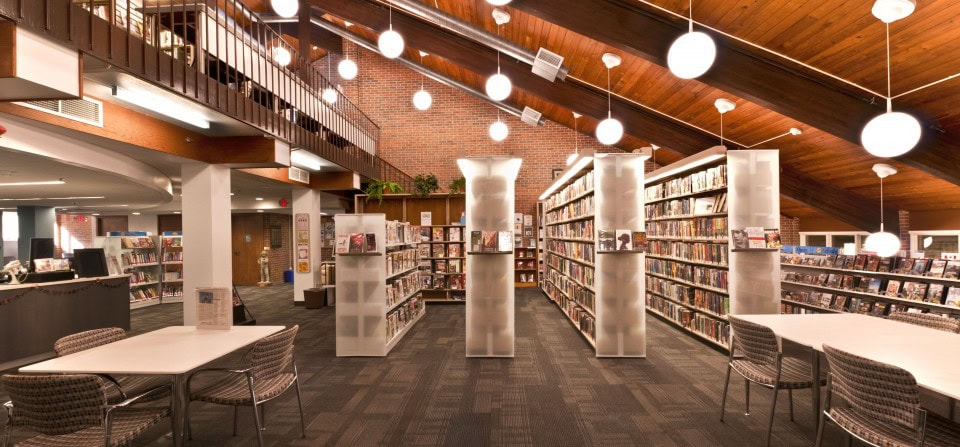 Modern library interior with high wooden ceilings, hanging globe lights, bookshelves filled with books, tables and chairs for reading, and a brick wall in the background.