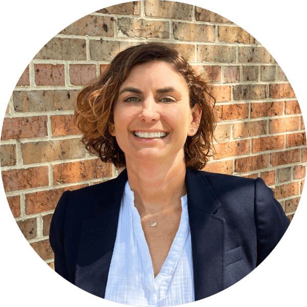 A woman with short, curly brown hair smiles at the camera. She is wearing a navy blazer over a white blouse, standing in front of a brick wall in sunlight.