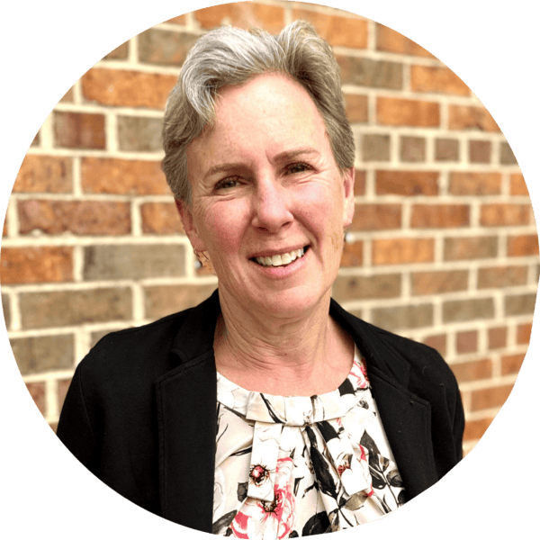 A smiling woman with short gray hair wearing a black blazer and floral blouse stands in front of a brick wall.